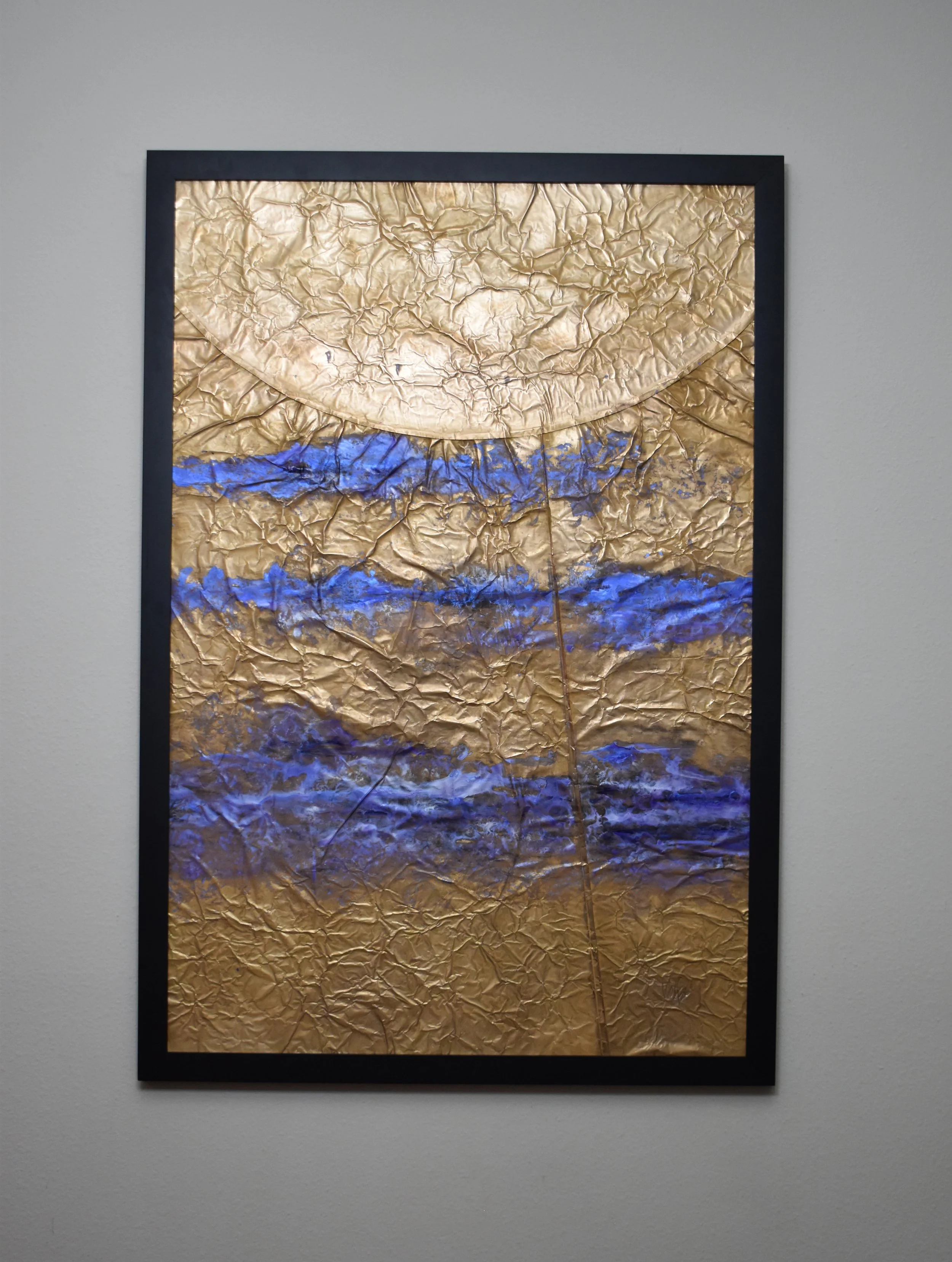 Textured gold-toned abstract artwork with horizontal bands of vivid blue and a large curved circular form extending across the top. Displayed framed on a white wall.