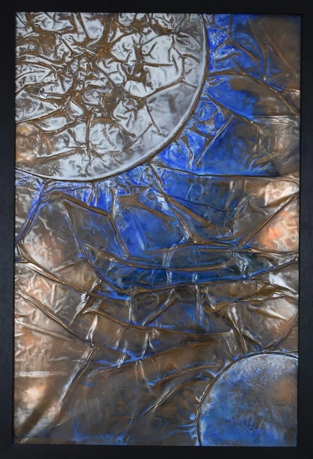 Planetary forms emerge through fractured currents of vivid blue and bronze. Shown close up in a black frame.