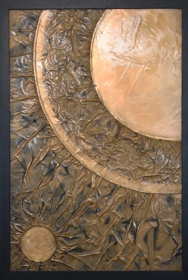 Golden planetary forms overlap within a dark metallic field. Shown close up in a black frame.