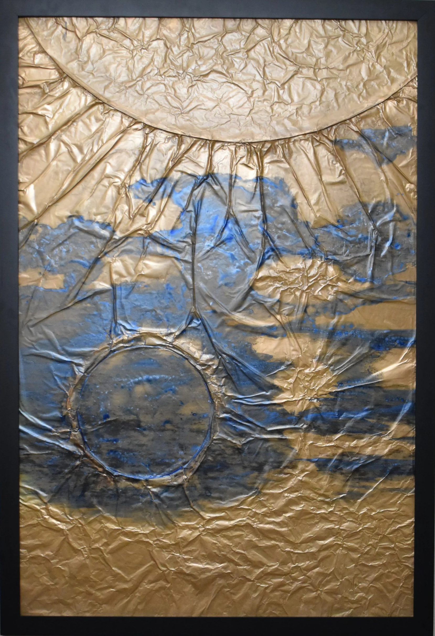 Textured gold-toned abstract artwork with blue and black markings, featuring a large curved form above and a dark circular form below. Shown close up in a black frame.