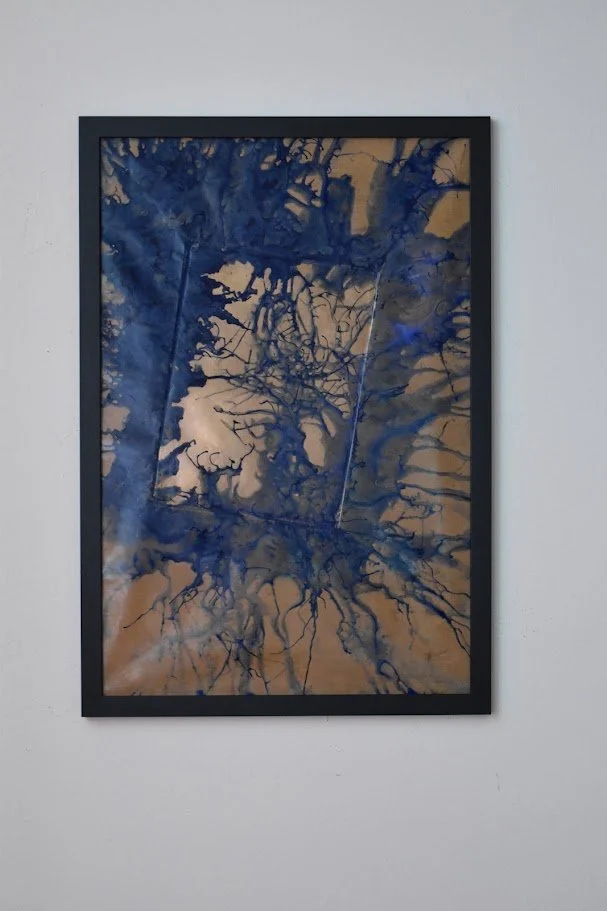 Deep blue currents spread across a warm metallic field. Displayed framed on a white wall.