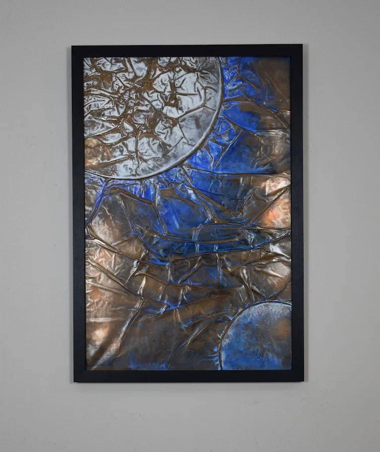 Planetary forms emerge through fractured currents of vivid blue and bronze. Displayed framed on a white wall.