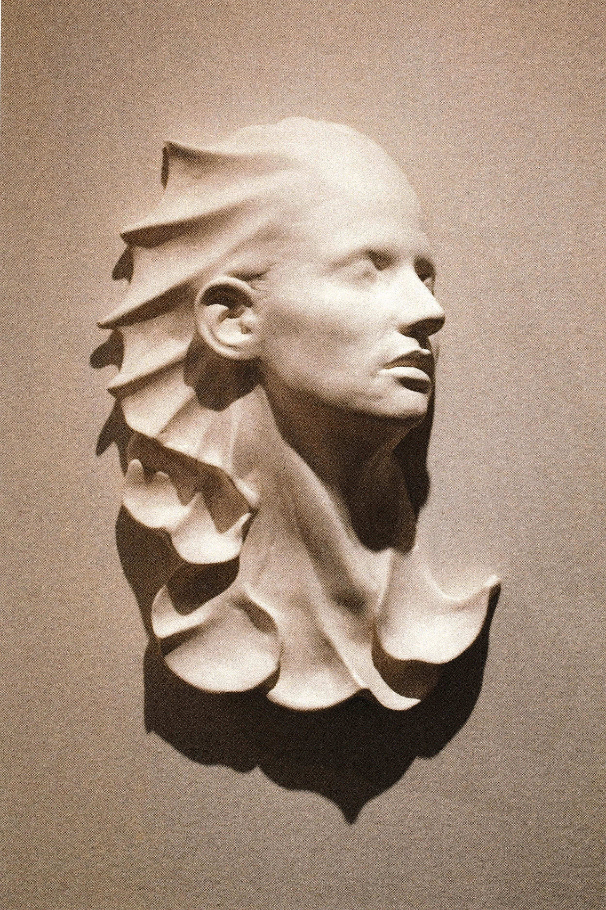 Wall-mounted life-mask-derived ceramic sculpture with ribbon-like forms radiating behind the head like windswept hair or unfolding petals. Aquatic creature looking.