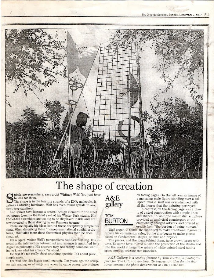 A black and white photograph of an outdoor scene showing a person standing behind a metal sculpture called 'The Shape of Creation', with trees and sky in the background.