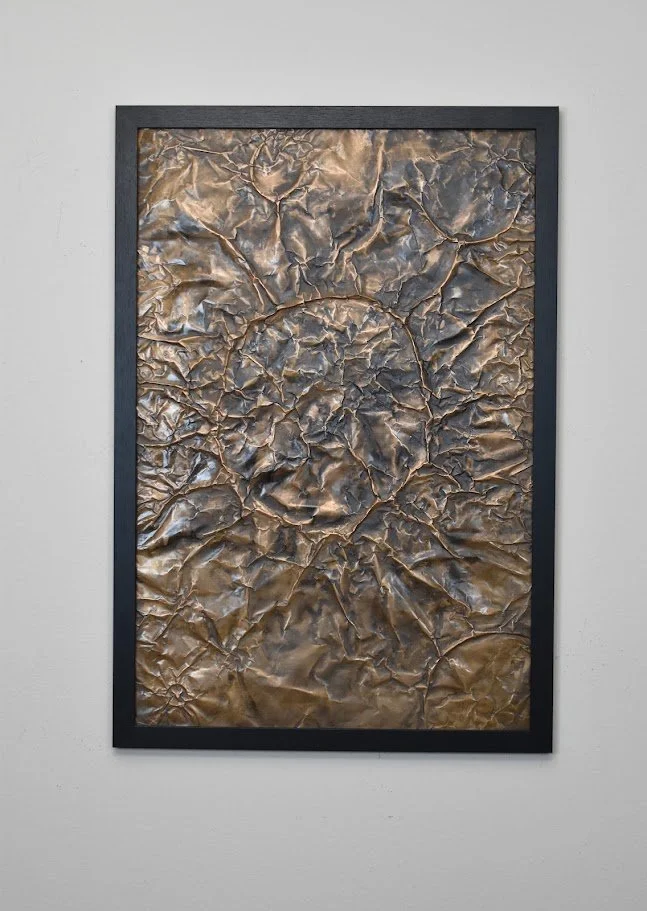 A fractured metallic surface in shifting bronze and charcoal tones. Displayed framed on a white wall.