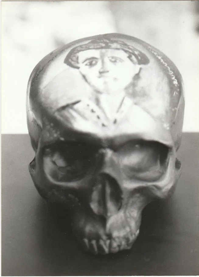 A black and white close-up view of a skull sculpture resting on a pedestal, shown from the front with deeply recessed eye sockets and a smooth, rounded cranium. The forehead contains a re-created "The Bullfighter" Picasso painting.