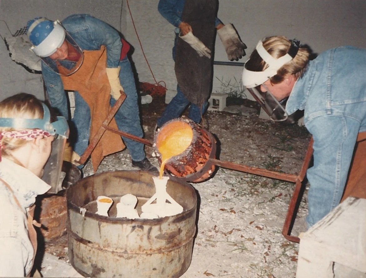 A group of artists wearing protective face shields and heat-resistant gear pour molten metal from a crucible into sculptural molds during a bronze casting process. Whitney Wolf II is the man on the right.