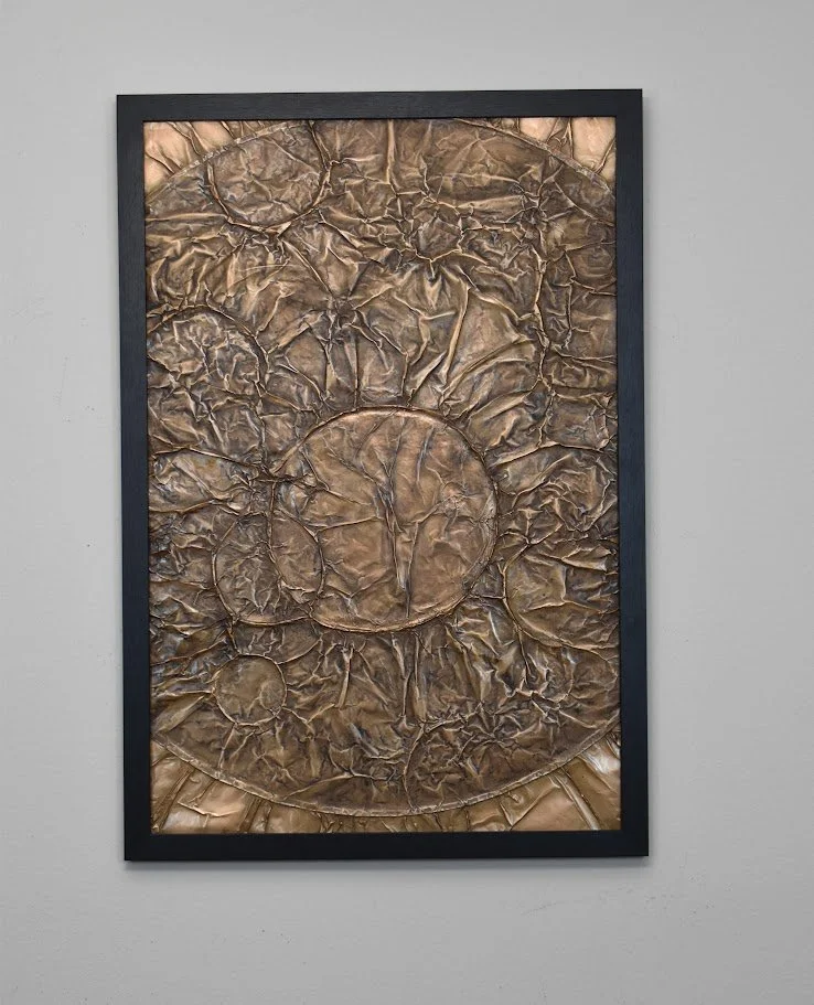 Circular forms emerge within a fractured bronze field. Displayed framed on a white wall.