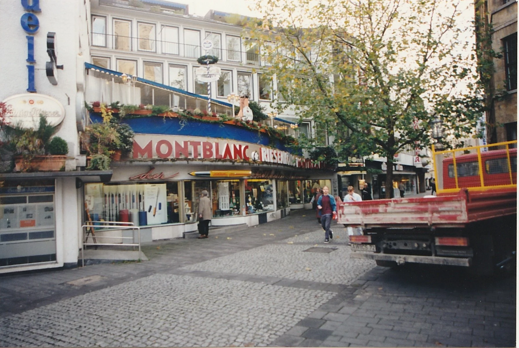 Busy city square with cafés, shops, and pedestrians beneath trees.