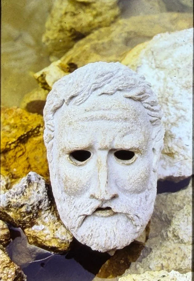 White sculpted mask depicting a bearded classical male figure with hollow eyes, resting among rocks in the ocean. The mask has a solemn expression and evokes ancient Greek sculptures.