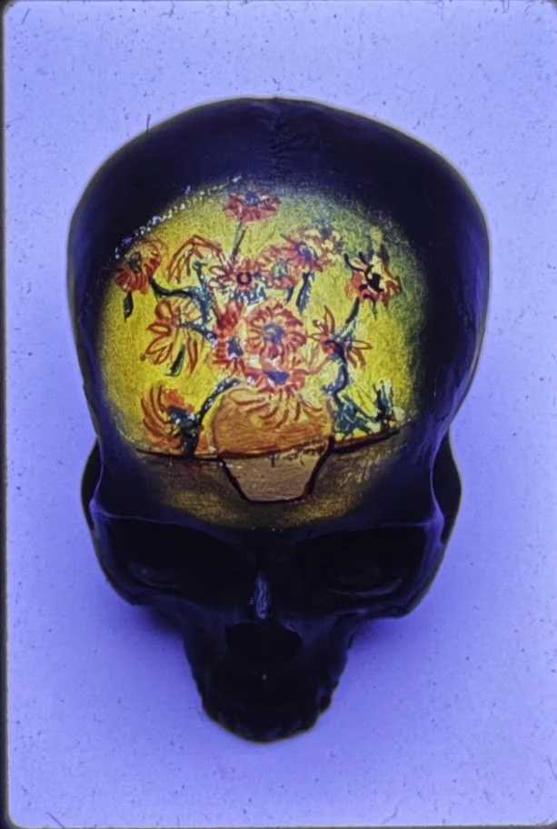 A full color close-up view of a skull sculpture resting on a pedestal, shown from the front with deeply recessed eye sockets and a smooth, rounded cranium. The forehead contains a re-created the "Sunflowers" Picasso painting.