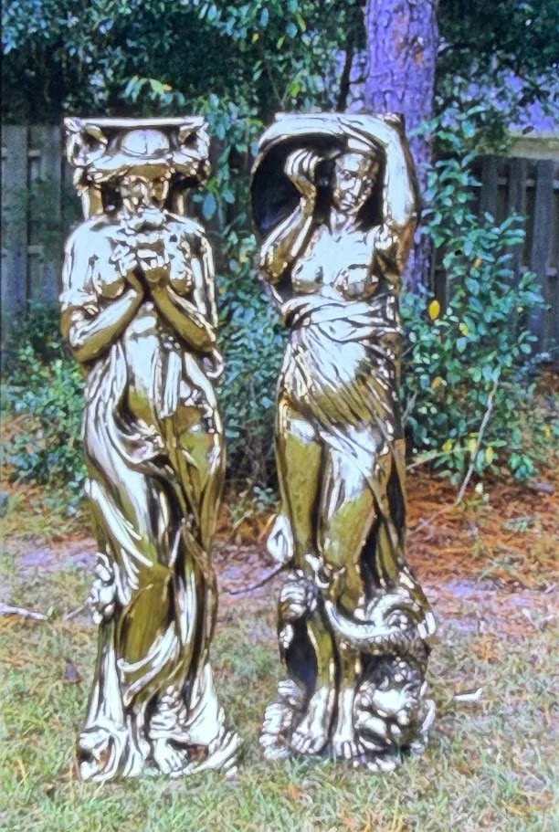 Pair of classical female figures standing outdoors, draped in flowing garments and posed beside decorative architectural elements.