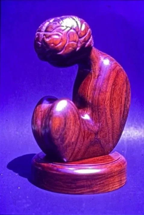Rounded wooden sculpture of a seated figure with knees drawn toward the body.