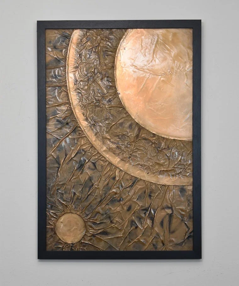 Golden planetary forms overlap within a dark metallic field. Displayed framed on a white wall.