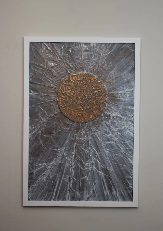A copper orb radiates across a silvery metallic field. Displayed framed on a white wall.