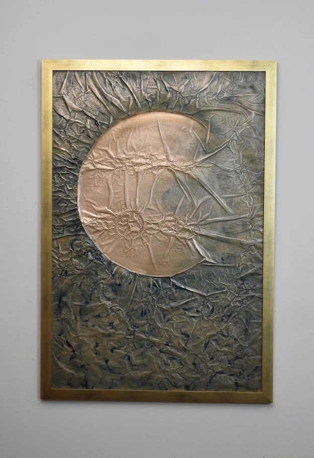 A copper orb emerges from a dark, textured metallic field. Displayed framed on a white wall.