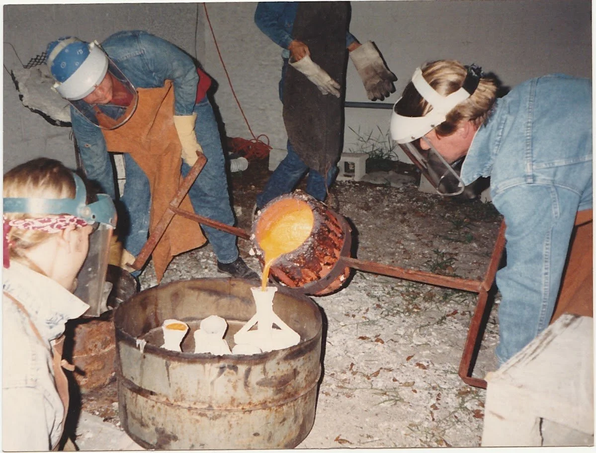 Whitney Wolf II pouring bronze into molds with the help of three students. Everyone is wearing protective gear and face shields.