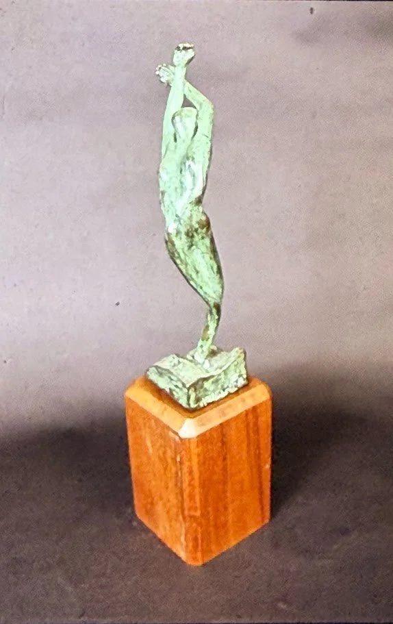Green-patinated bronze dancer rising from a warm wooden pedestal, the body stretching upward in a fluid gesture reminiscent of a leaf unfurling in the wind.