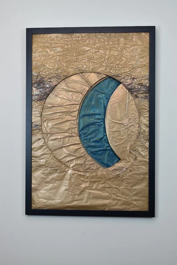 Interlocking golden and blue forms create a planetary eclipse. Displayed framed on a white wall.