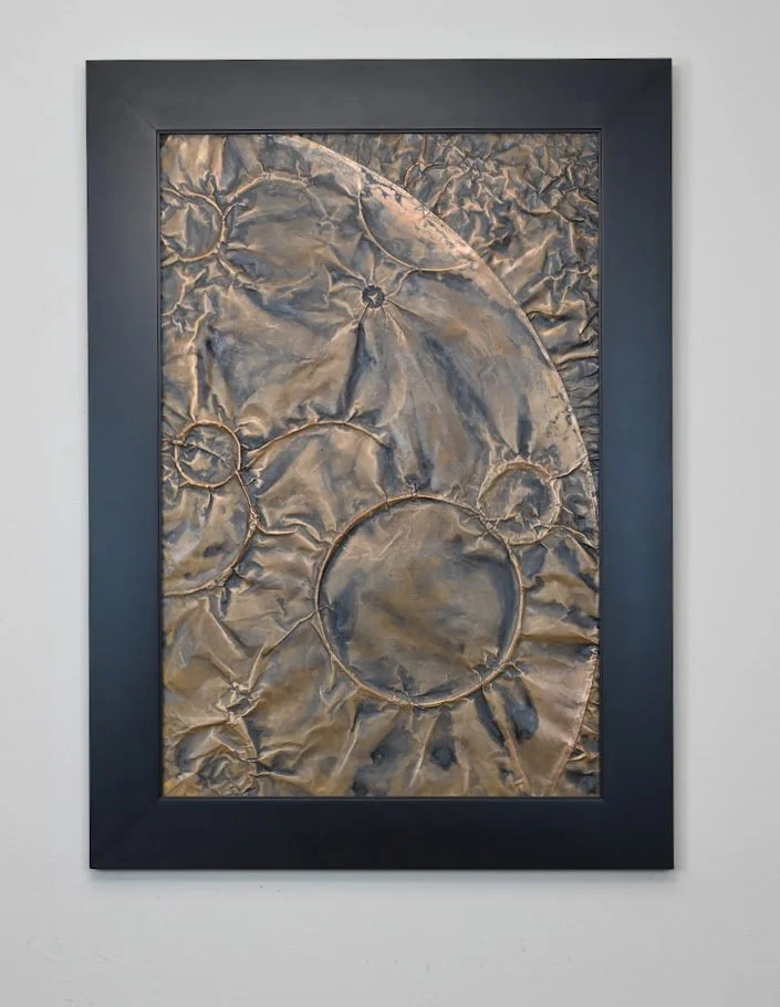 Circular forms drift across a fractured metallic field. Displayed framed on a white wall.