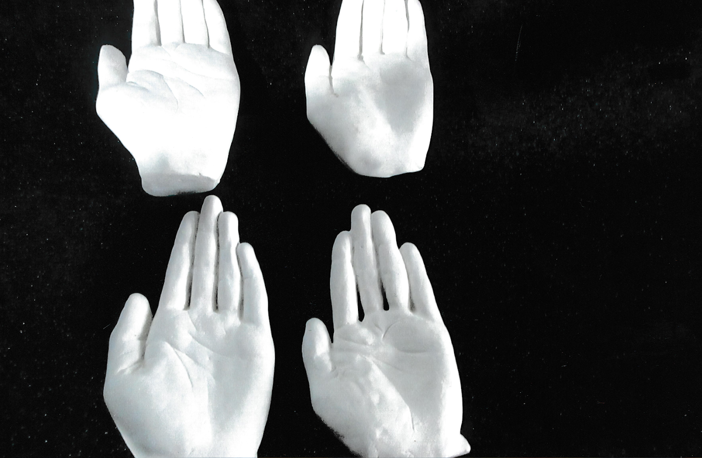 Four white plaster casts of human hands are arranged ina  grid against a black background. The open palms appear suspended in space.