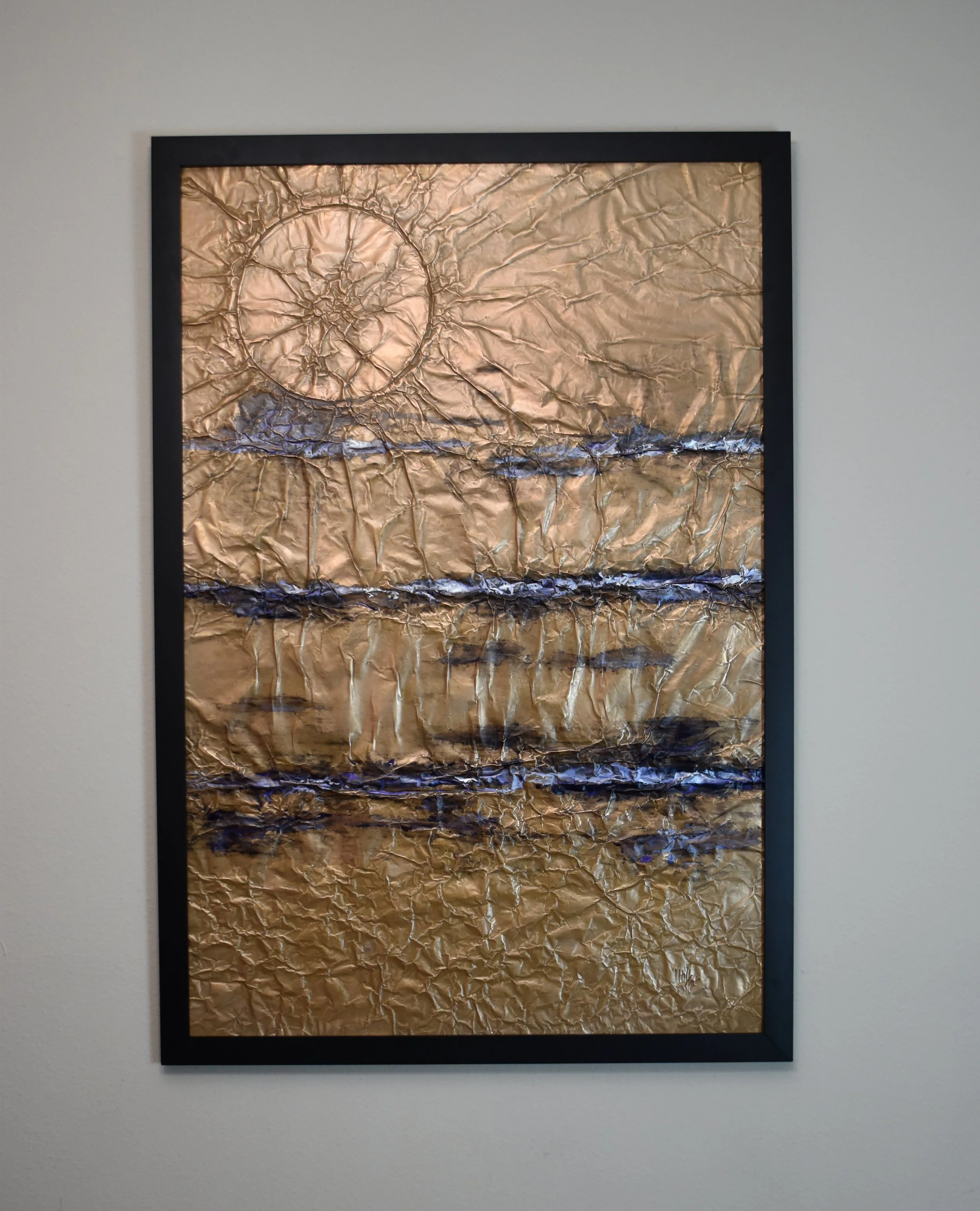Textured gold-toned abstract artwork with horizontal blue bands and a large circular form in the upper left. Displayed on a white wall.