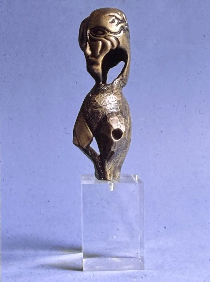 Elongated bronze figure with stylized facial features and an open mouth, displayed on a clear acrylic pedestal. The narrow body tapers toward a small base.