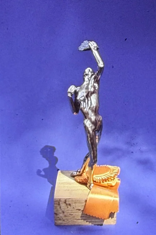Small bronze sculpture of an elongated female figure standing atop a wooden base, raising a hand mirror above her head while the opposite arm bends gracefully near her chest. The slender body is modeled with flowing, organic contours that catch the l