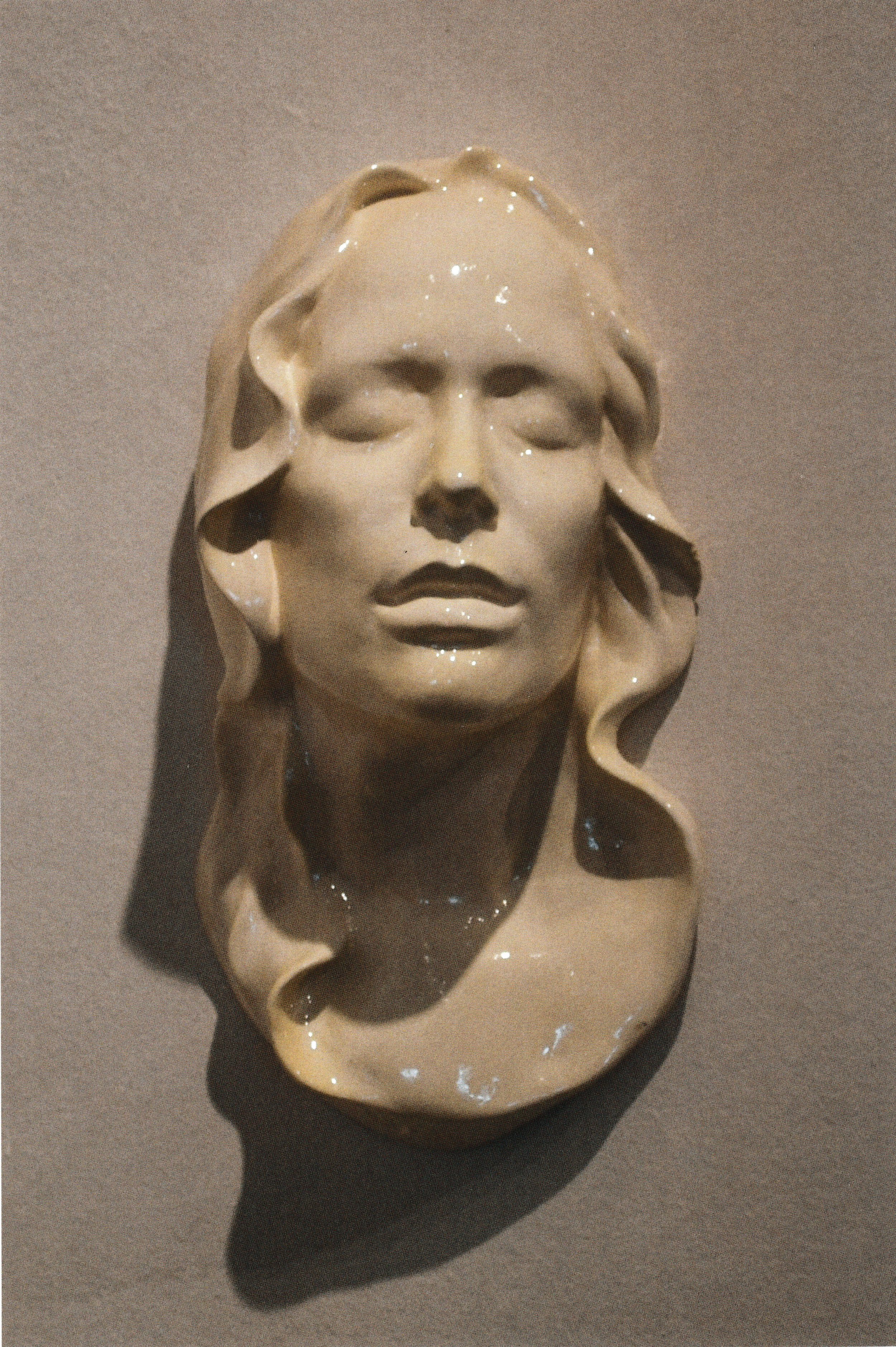 Wall-mounted ceramic life mask with closed eyes and softly curling hair framing the face.