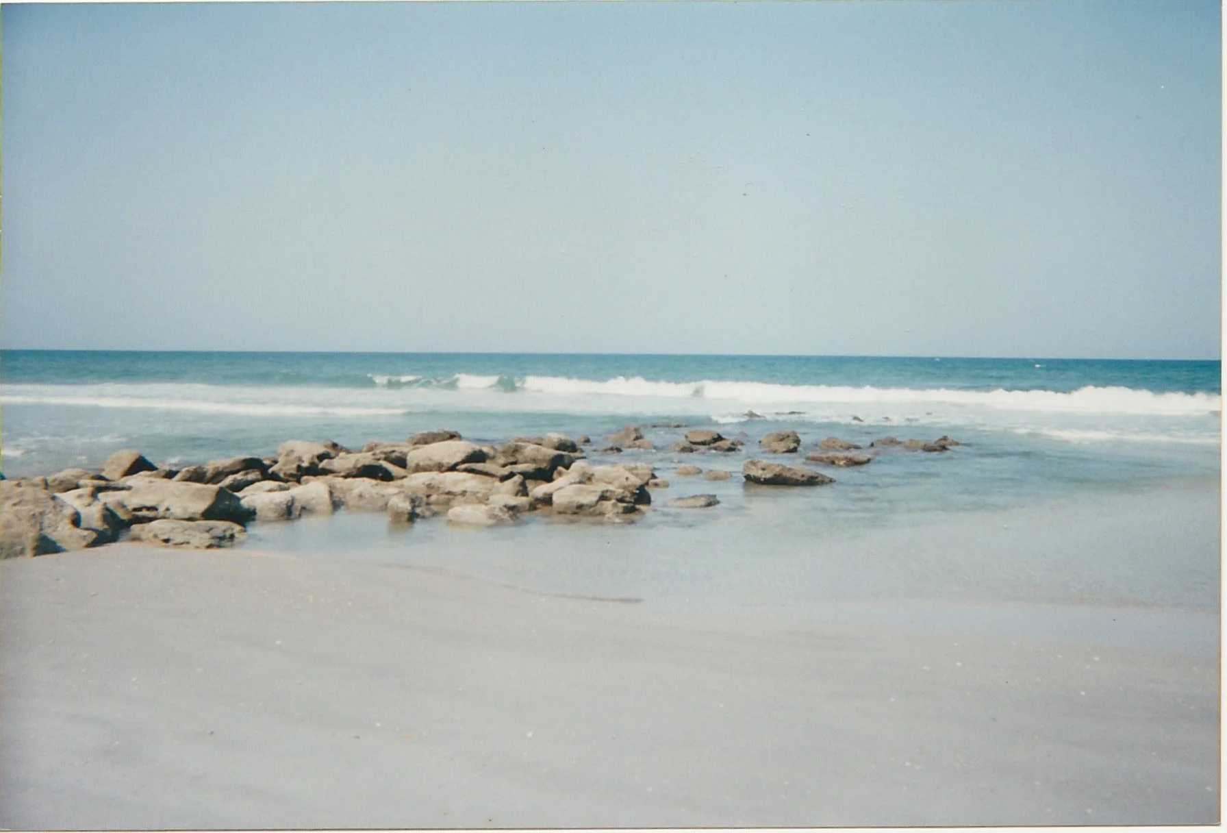 Rocky shoreline meeting calm ocean waves beneath a pale blue sky.