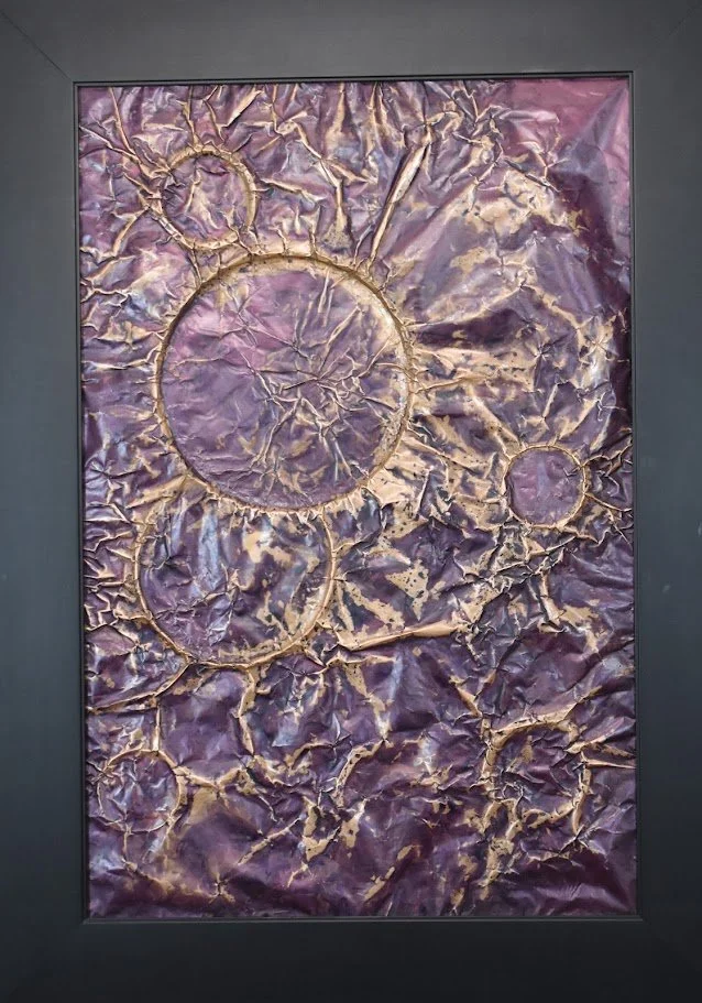 Clusters of circular forms emerge through layers of violet, lavender, and gold, resembling distant worlds suspended within a turbulent field. Shown close up in a black frame.