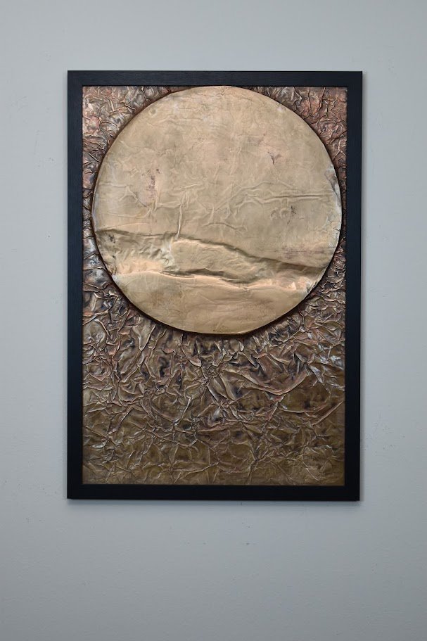 A golden orb floats within a textured bronze field. Displayed framed on a white wall.