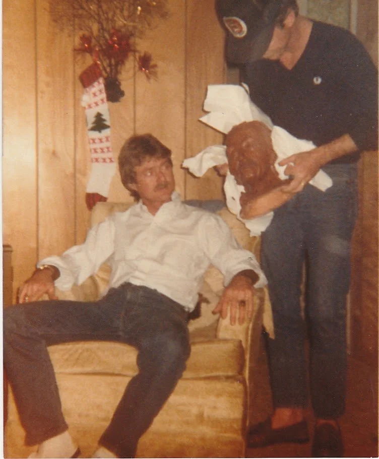 Whitney Wolf II is sitting in a chair during the holiday season, being presented with a clay bust carving by a friend. Circa 1979