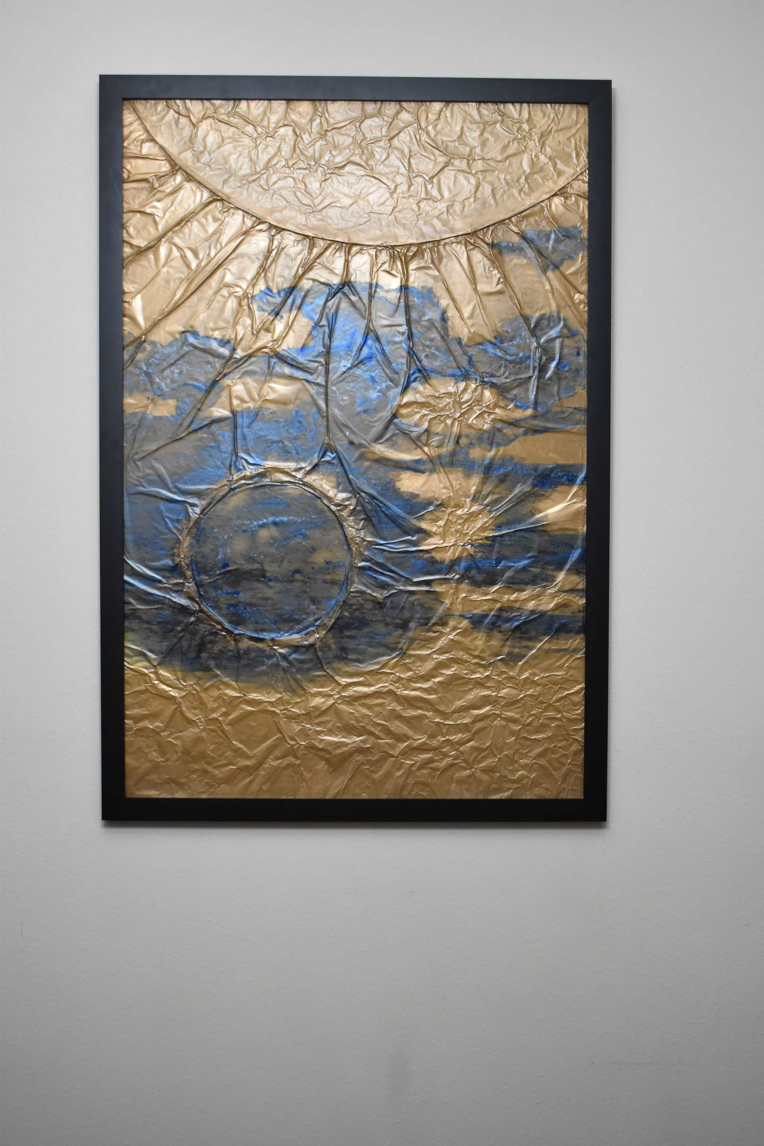 Textured gold-toned abstract artwork with blue and black markings, featuring a large curved form above and a dark circular form below. Displayed framed on a white wall.