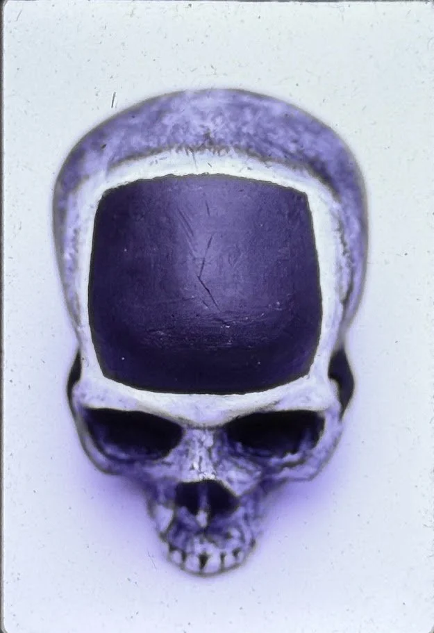 A black and white close-up view of a skull sculpture resting on a pedestal, shown from the front with deeply recessed eye sockets and a smooth, rounded cranium. The forehead contains what looks like a black television screen.