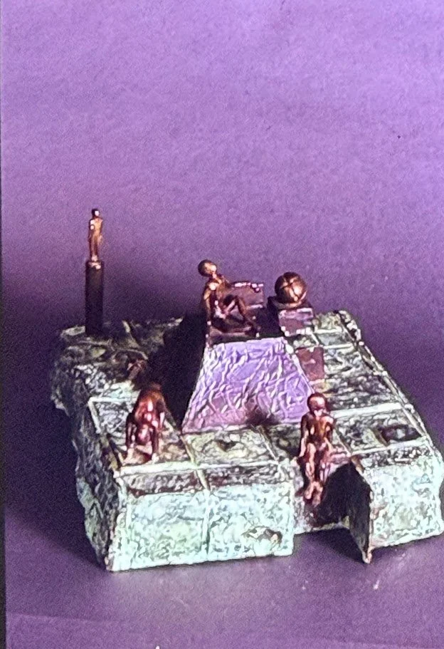 Small bronze environment resembling a walled courtyard. Human figures occupy different levels of the structure surrounding a raised central form.