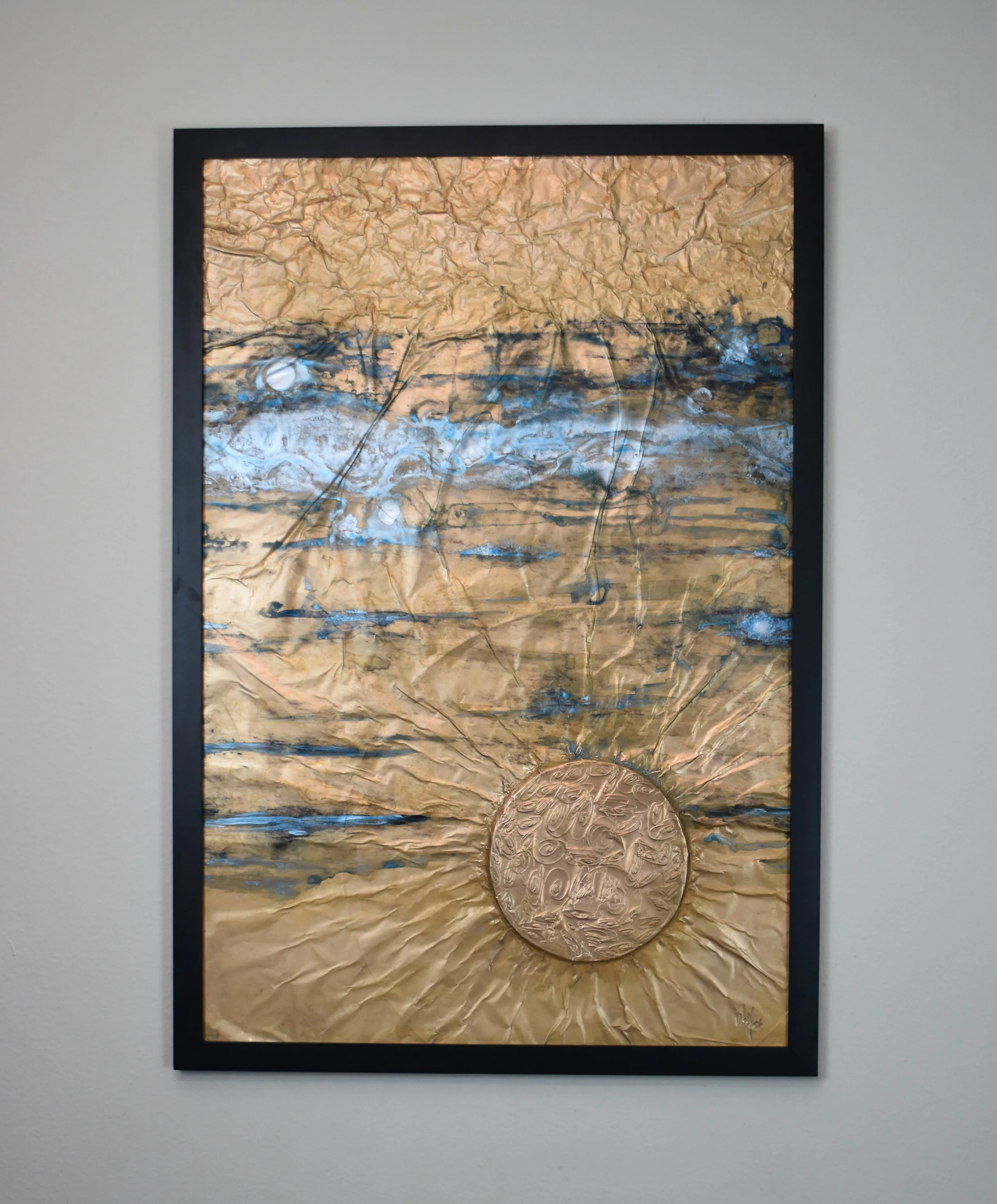 Textured gold-toned abstract artwork with blue and black horizontal markings and a raised circular form radiating outward near the bottom. Displayed framed on a white wall.