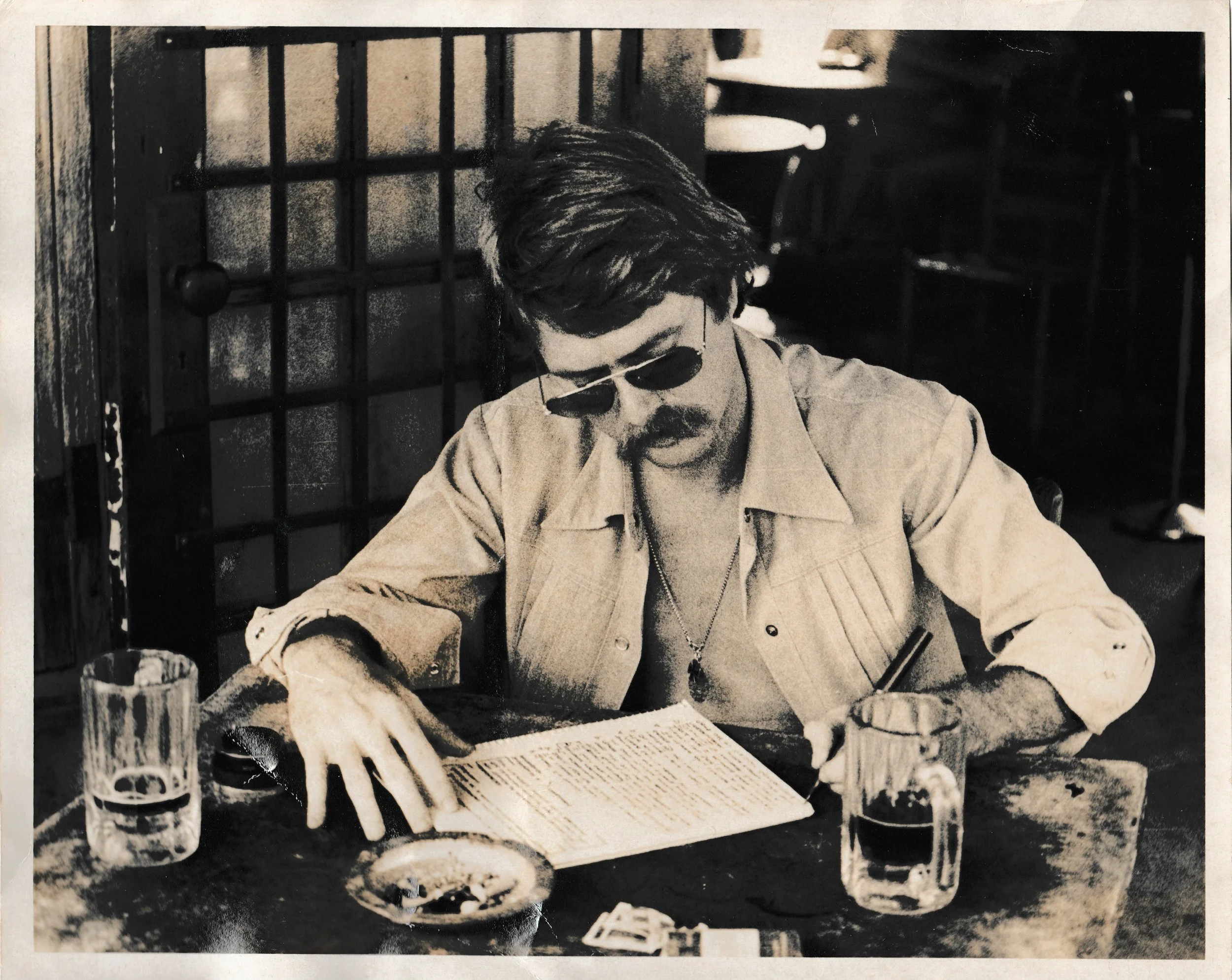 Black and white photo of a man with sunglasses sitting at a table with newspapers, pen, and glasses. There are drinks on the table, and shelves with plates and cups in the background.