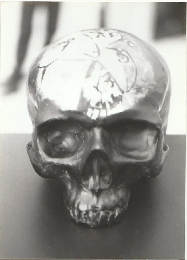 A black and white close-up view of a skull sculpture resting on a pedestal, shown from the front with deeply recessed eye sockets and a smooth, rounded cranium. The forehead contains a re-created Picasso painting.