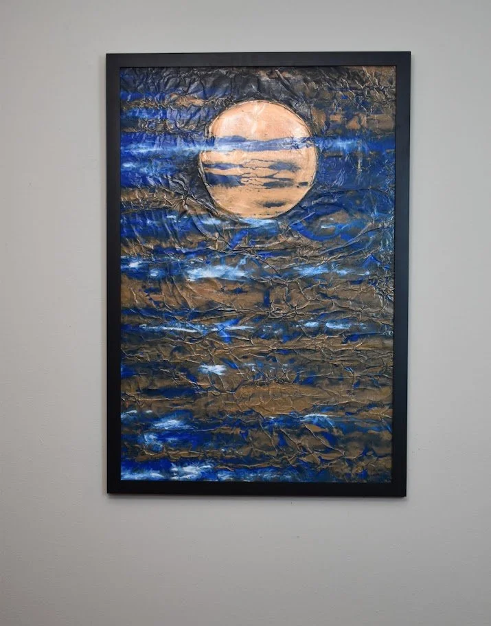 A golden orb floats within turbulent currents of deep blue. Displayed framed on a white wall.