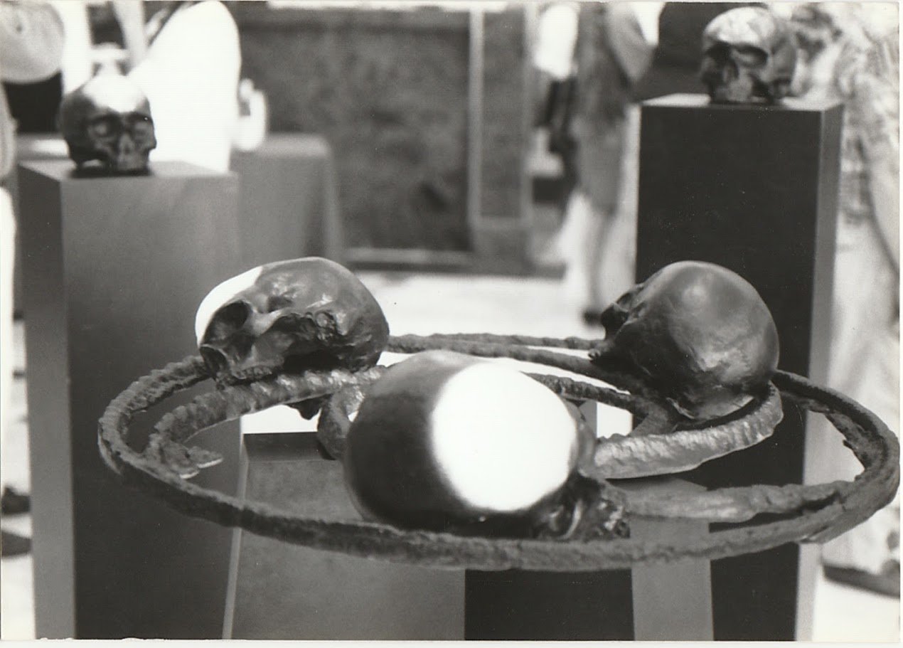 A suspended circular arrangement of skulls serves as the focal point of an installation composed of individual skull sculptures displayed on pedestals.