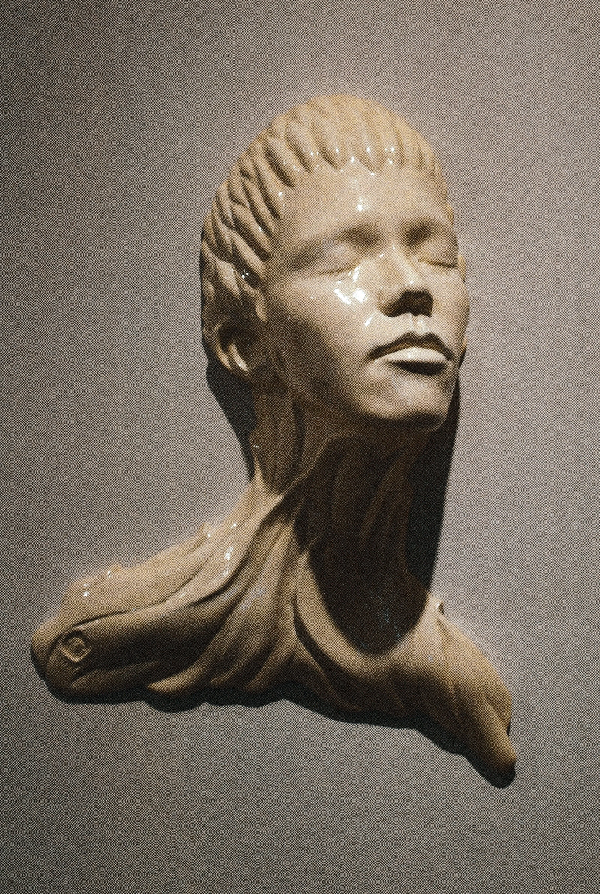 Wall-mounted ceramic life mask bust with eyes closed and fabric-like forms cascading from the shoulders.