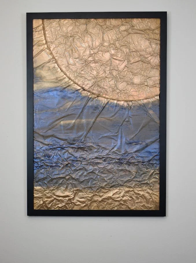 A massive golden form curves into view above layers of deep blue and metallic earth tones. Displayed framed on a black wall.