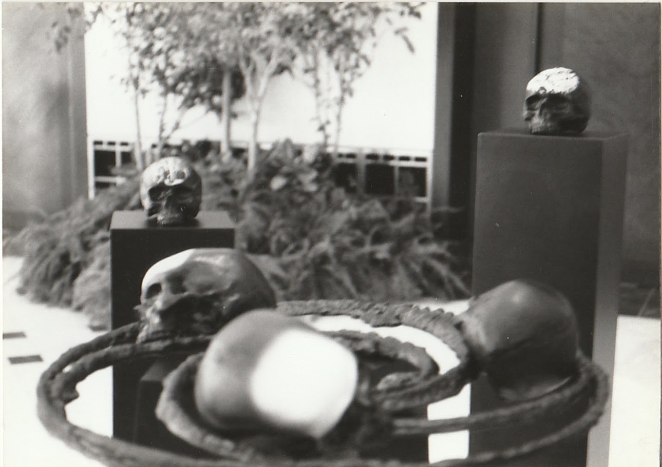 A circular sculptural structure supports multiple skull forms above a pedestal, surrounded by additional skull sculptures positioned throughout the exhibition space.