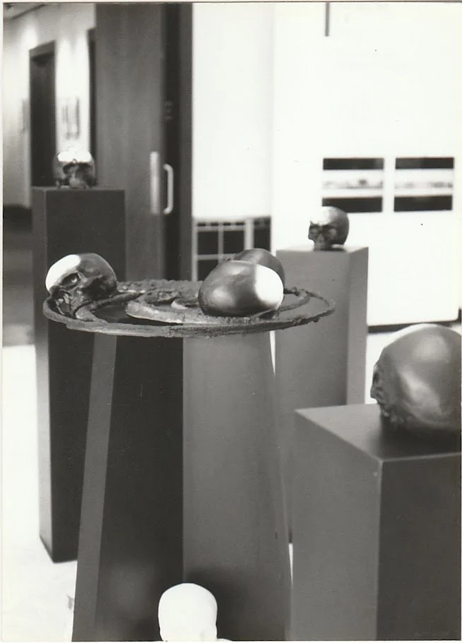 Black-and-white gallery photograph showing skull sculptures displayed on tall pedestals. A circular sculptural arrangement in the center connects several skull forms, creating an installation that explores themes of mortality, memory, and human exist
