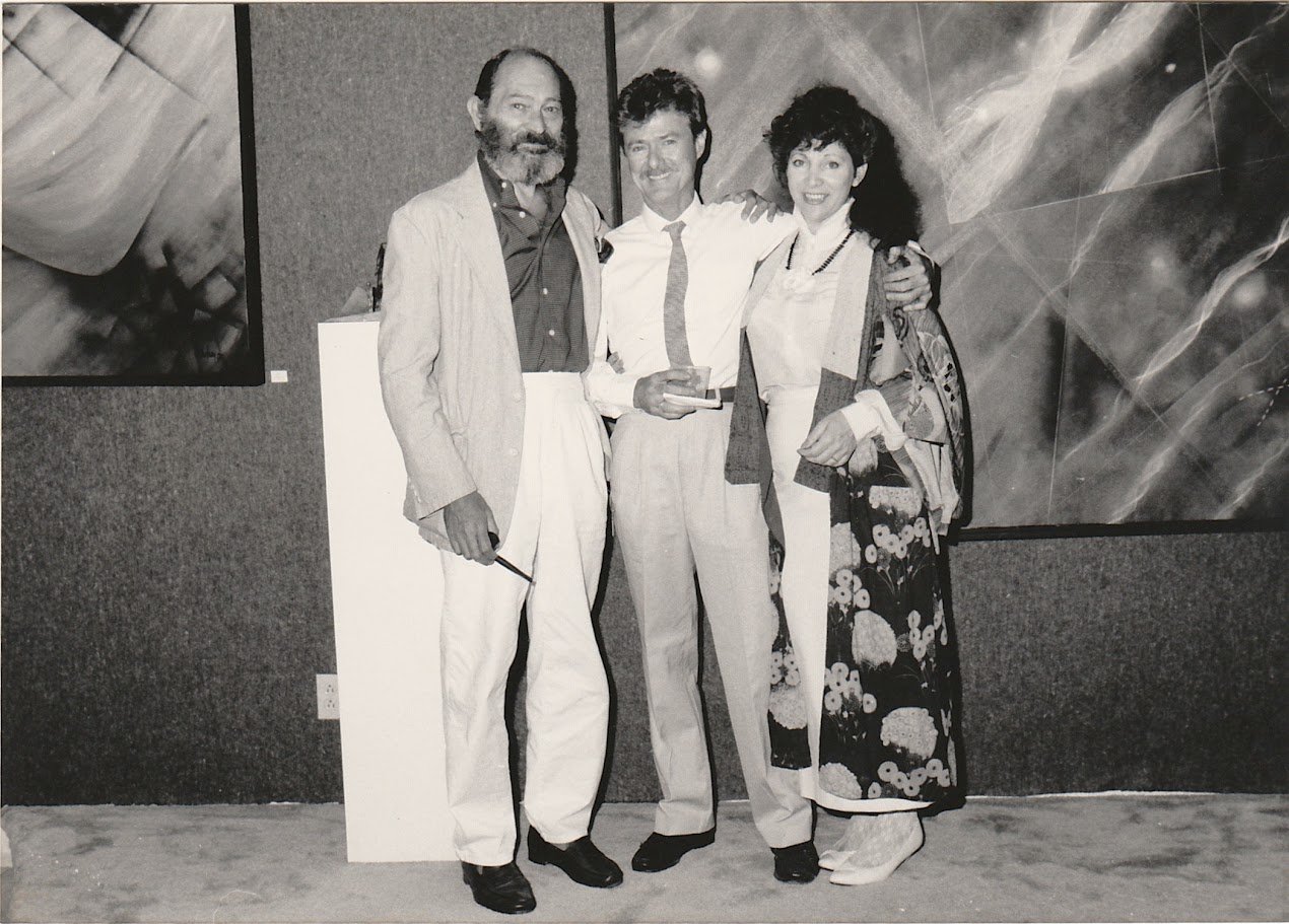 Whitney Wolf II featured in a black-and-white photo, standing in front of giant paintings and smiling. Wolf is in between his philosophy mentor (right) and a woman dressed in a Kimono. Est 1980s