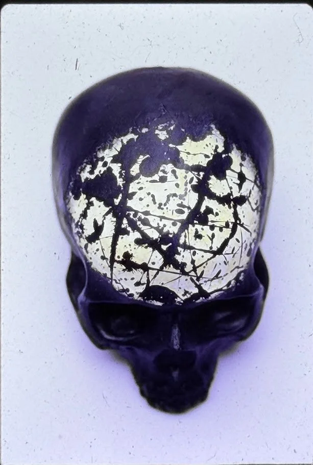 A black and white close-up view of a skull sculpture resting on a pedestal, shown from the front with deeply recessed eye sockets and a smooth, rounded cranium. The forehead contains a re-created the "Black and White" painting by Jackson Pollock. 