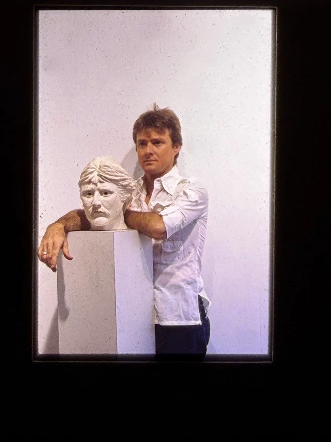 Digitized slide portrait photograph of artist Whitney Wolf II, standing beside a sculpted portrait bust. He rests his arms on the pedestal supporting the plaster head.