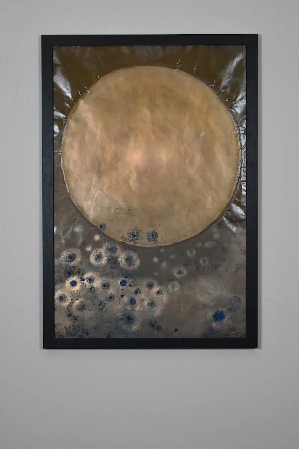 A textured metallic composition dominated by a large golden orb above a field of scattered blue and silver circular forms. Displayed framed on a white wall.