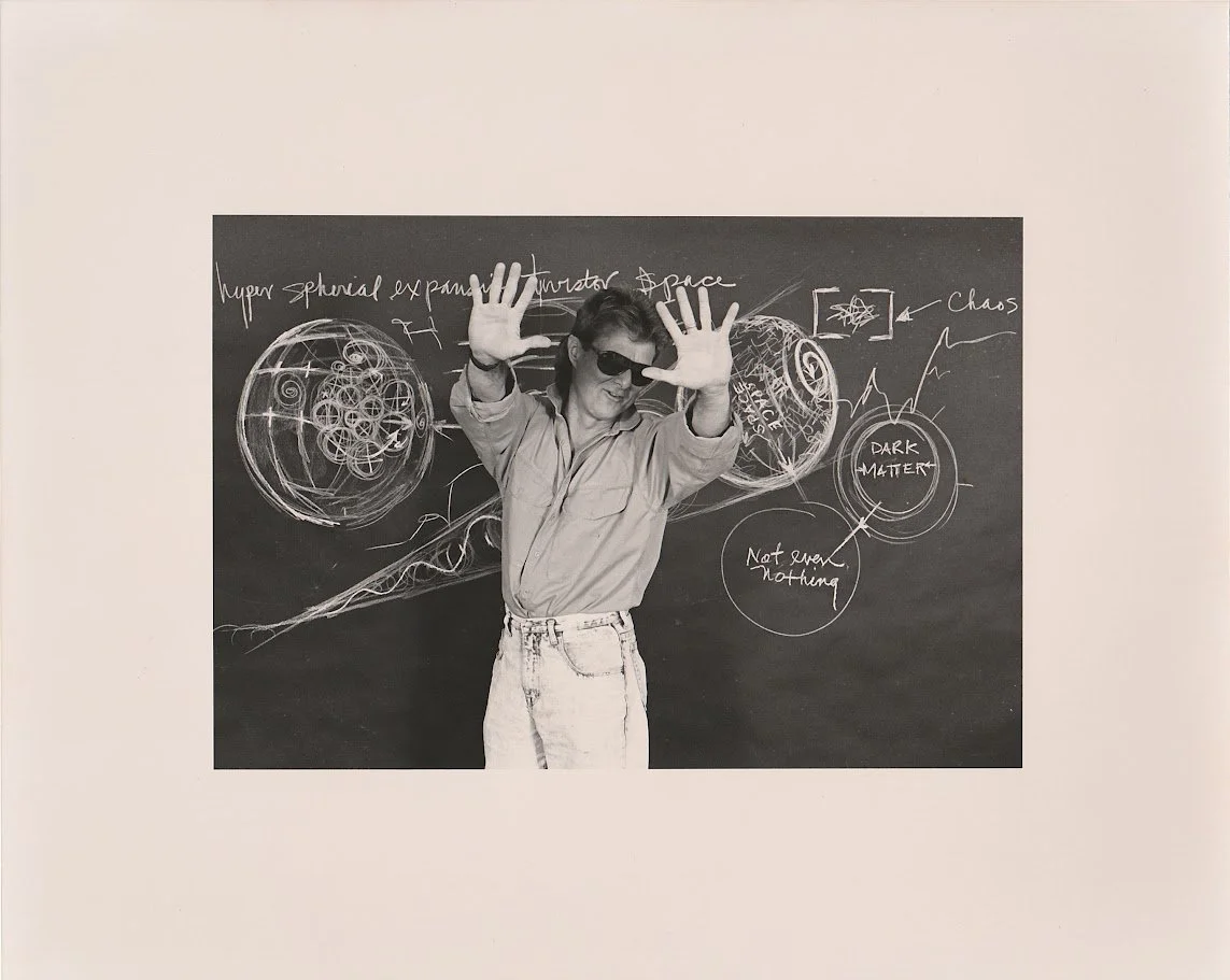 A black and white photo of Whitney Wolf in front of a chalk board. He is wearing acid wash jeans, a button down shirt that has been rolled up, and sunglasses. He is reaching both of his hands out, playfully depicting himself as not wanting any photog